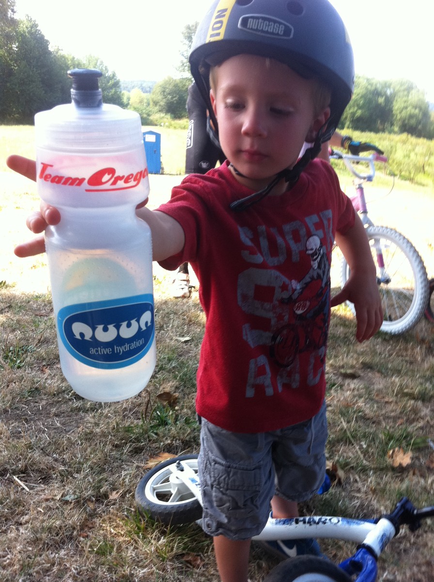 Preschool age child in a bike helmet, handing a water bottle toward the photographer