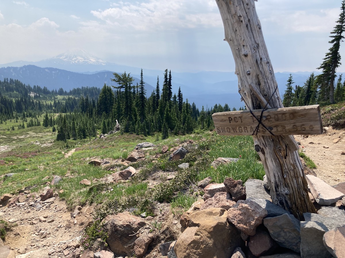 PCT Trail and Snowgrass trail junction sign on Snowgrass flats A heavily repaired trail junction sign and cairn in the foreground on a rocky alpine meadow with a stand of firs in the near distances. Mt. Adams on the horizon, its peak cloaked in haze and clouds