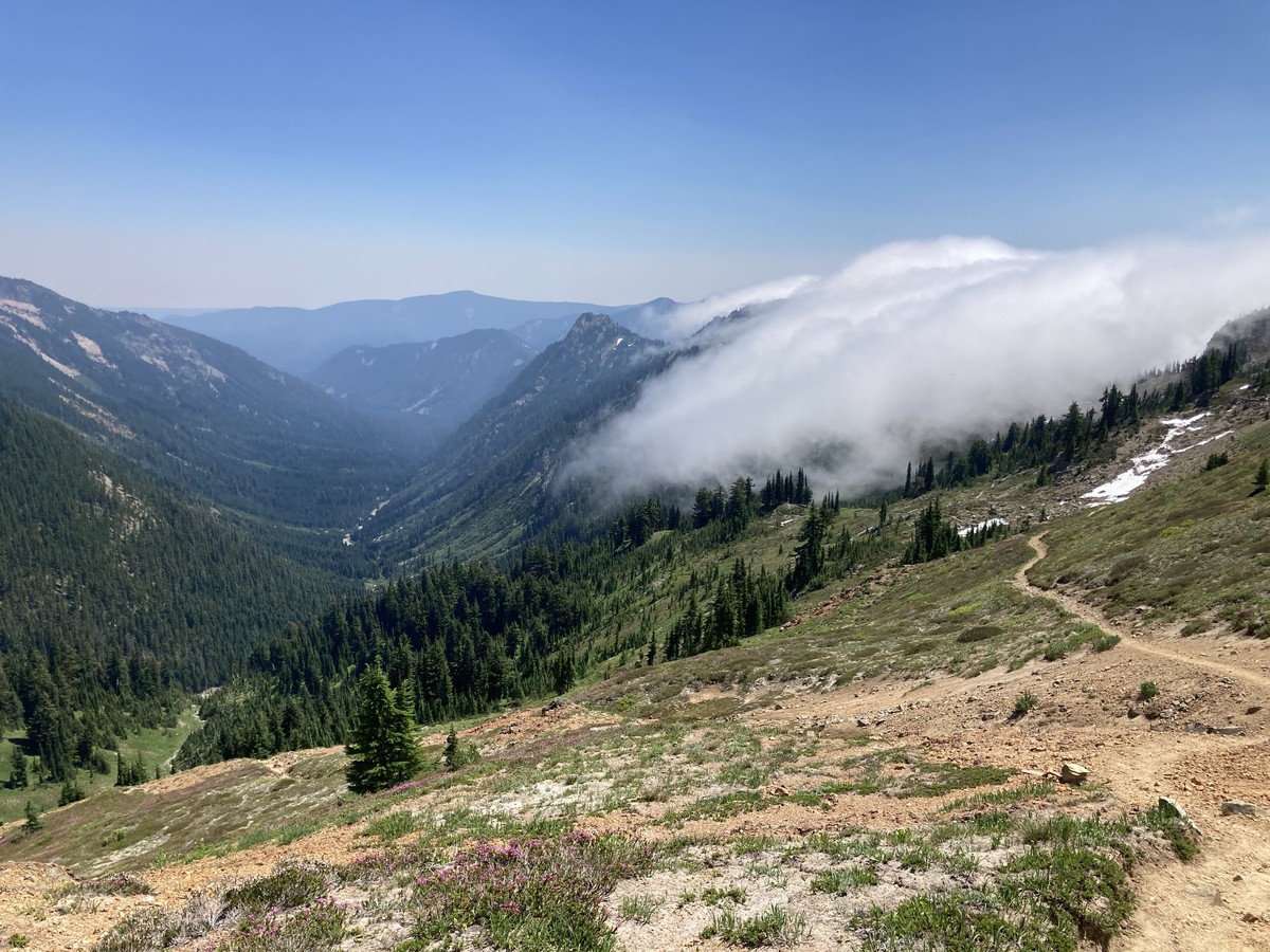 Cispus Pass descending into a foggy marine layer View from the top of a barren alpine pass looking down a u-shaped glacial valley toward forested mountains. On the right/west, a thick fog of marine clouds rolls over the ridge from the neighboring valley