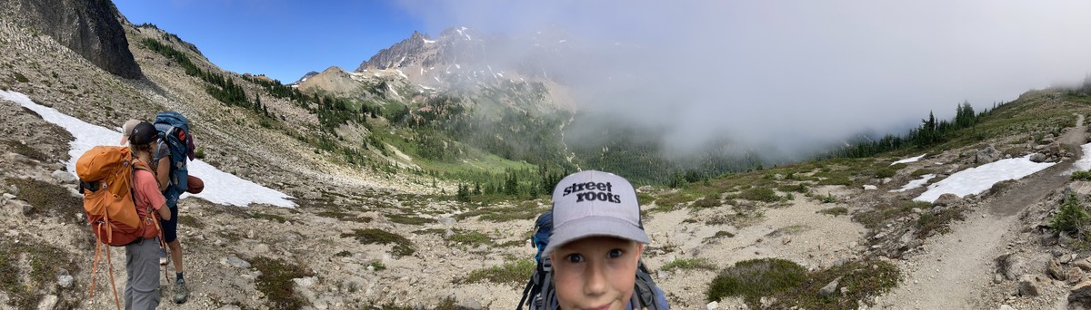 Cispus Pass, with a marine layer rolling in from the west Panorama of a high alpine valley with a young person in the foreground, looking into the camera, wearing a STREET ROOTS cap. We are just at the boundary where a layer of marine clouds are rolling into the valley, experienced here as a heavy fog to one side and clear sunlight on the other