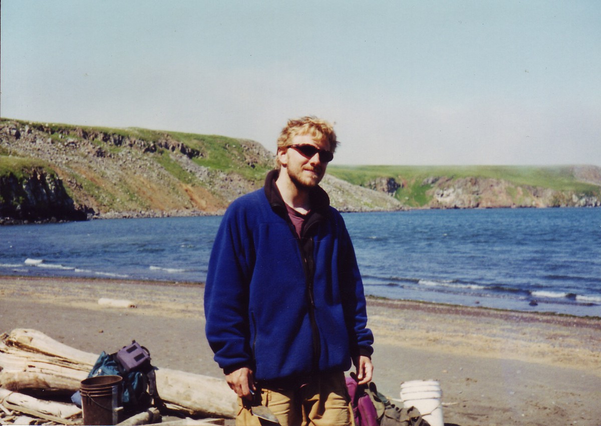 Nash Harbor, Nunivak Island, AK, 1996 Casual portrait of a disheveled young man with a beard and sunglasses, wearing a dingy fleece jacket, holding a mason’s trowel. Taken on a sunny day on beach in the subarctic, with green cliffs across a narrow bay in the background